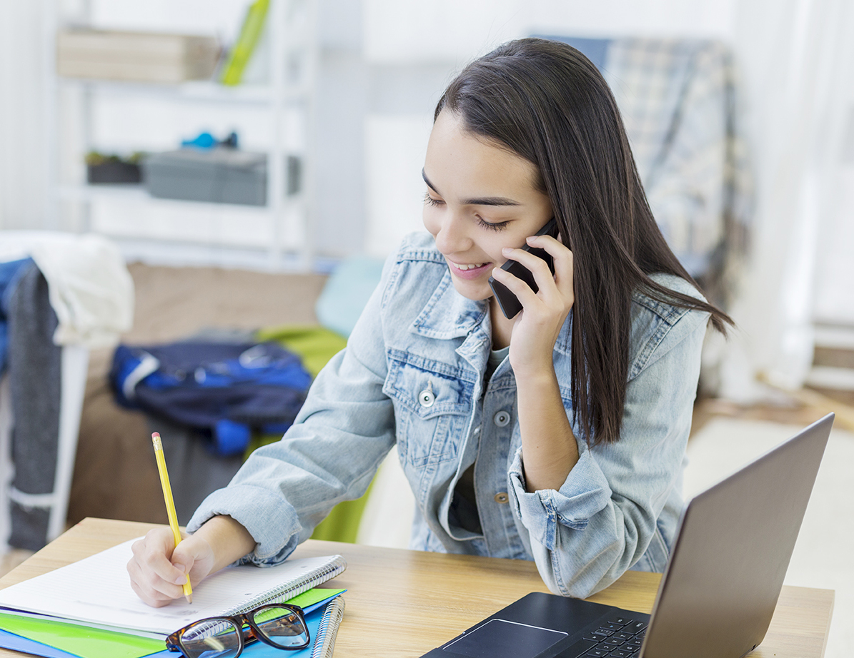 Pretty Hispanic college girl writes something in a notebook while talking on a smart phone. She is also using a laptop.